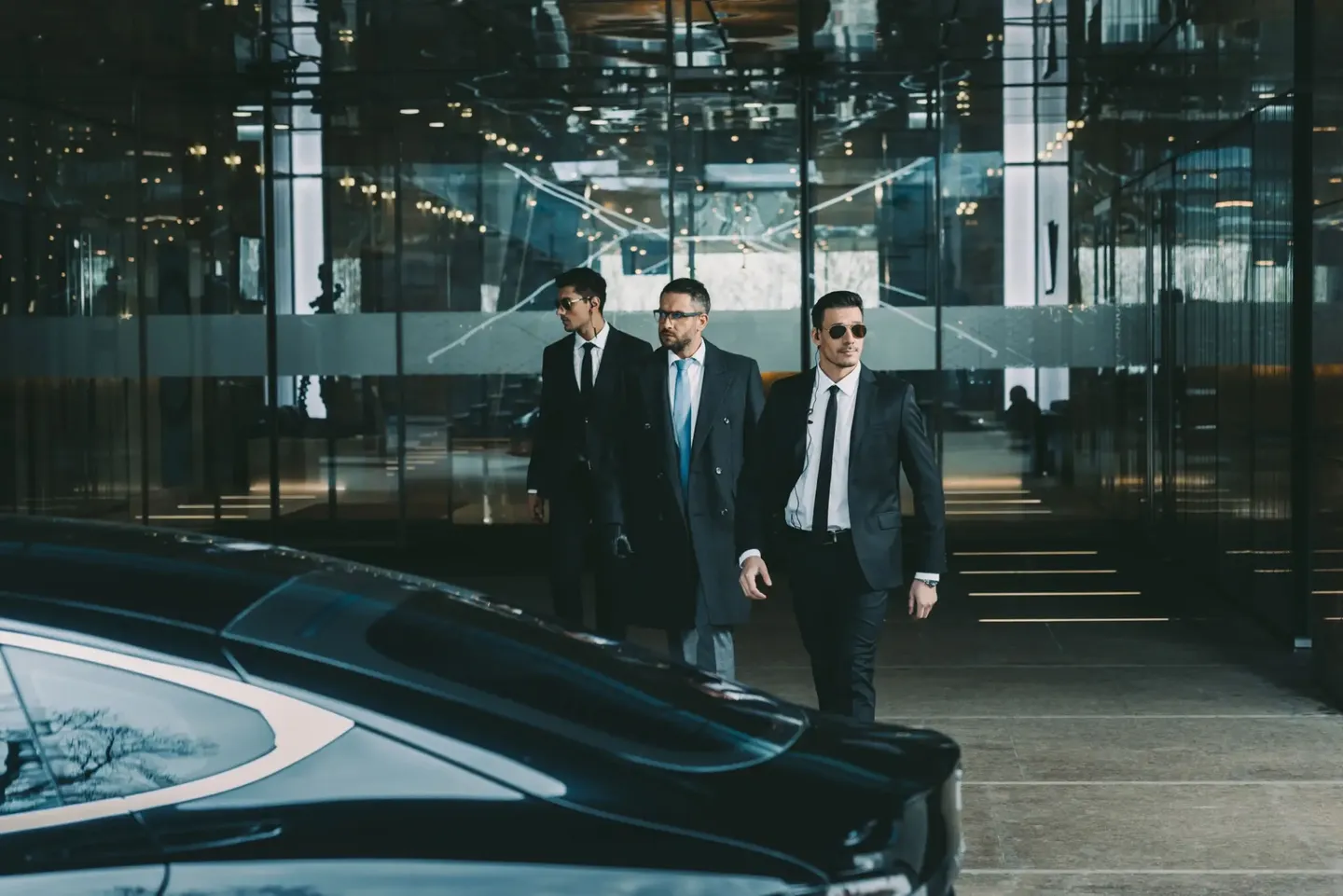 Three men in suits walking confidently near a luxury car.